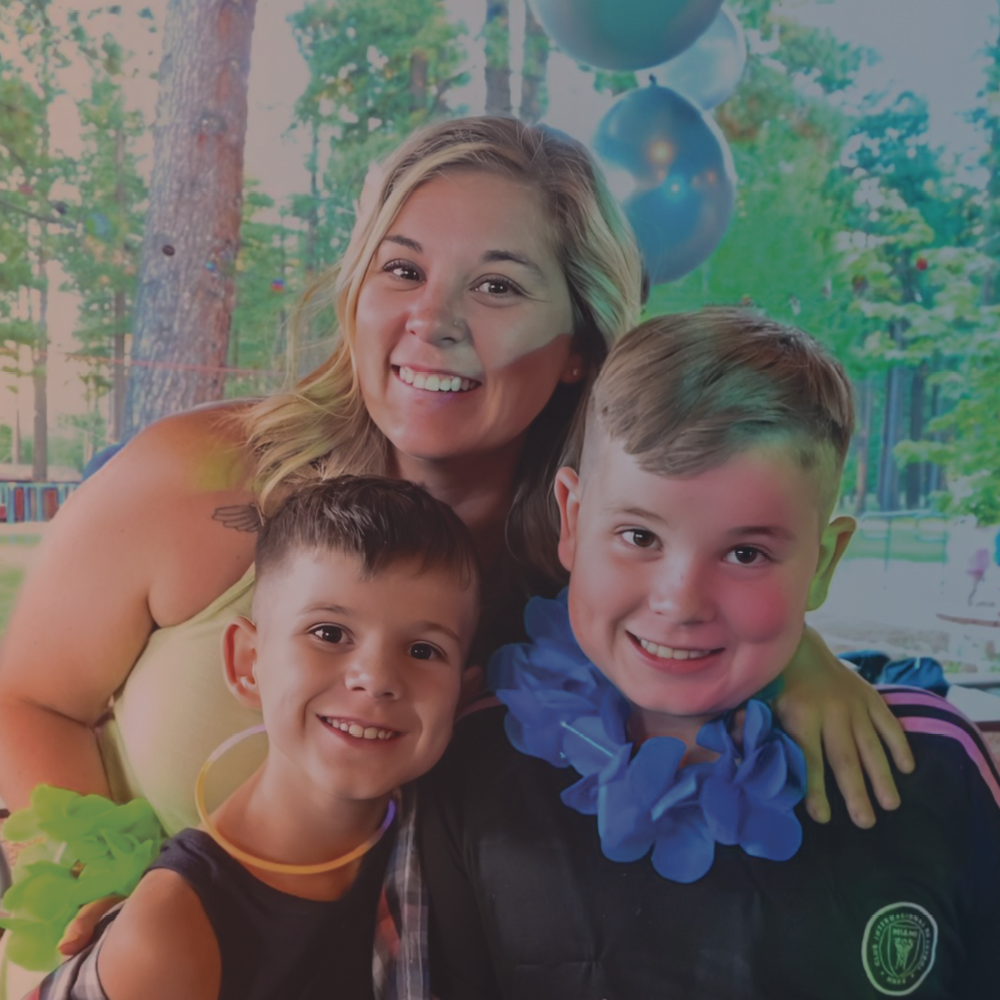 A young adult woman in a green top smiles with two young boys at a camp.