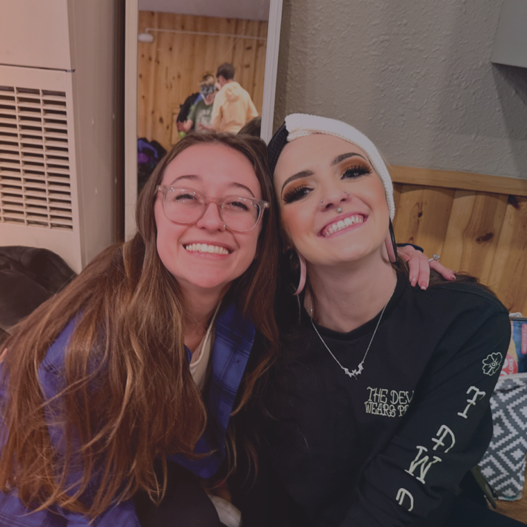 Two young girls smile and pose next to one another. The girl on the left is wearing a blue flannel, the girl on the right is wear a black long sleeve.