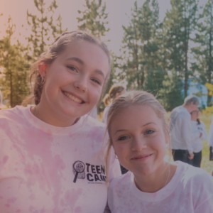 Two teenage girls smile together. They are wearing pink tye dyed shirts that read 2024 Teen Camp.